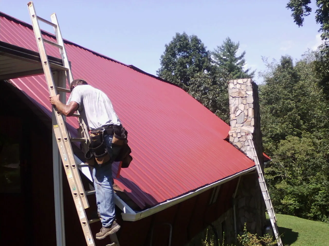 Roofer on a ladder beside a red metal roof and stone chimney — Soffit & Fascia Repair by Roofing Brothers in Sneedville